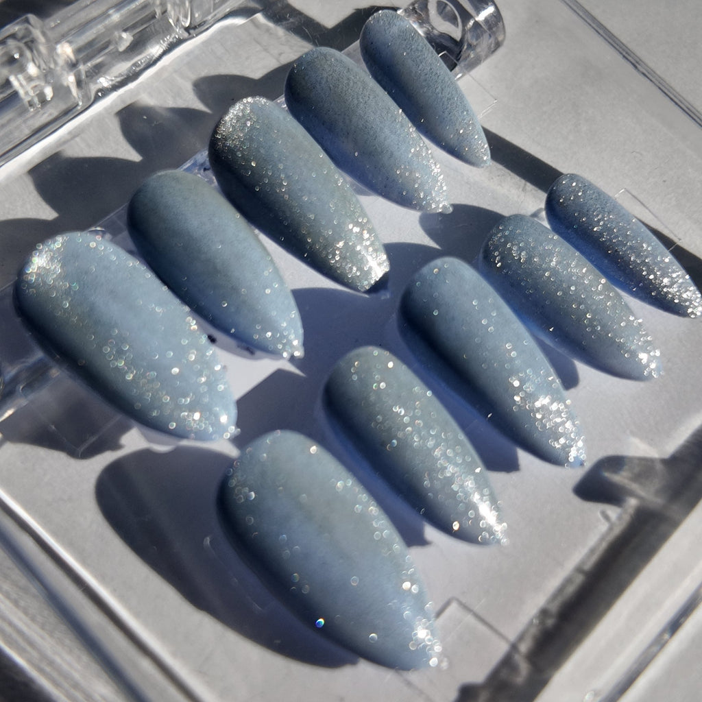 A set of press-on nails with a light blue glitter finish, displayed inside a clear plastic case on a reflective gray surface.