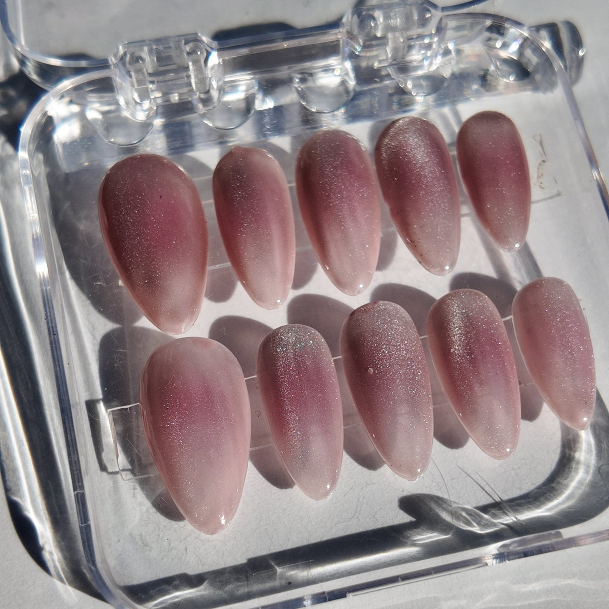 A set of ten false press-on nails in a clear plastic case, featuring a glossy pink gradient design with a subtle glittery texture.