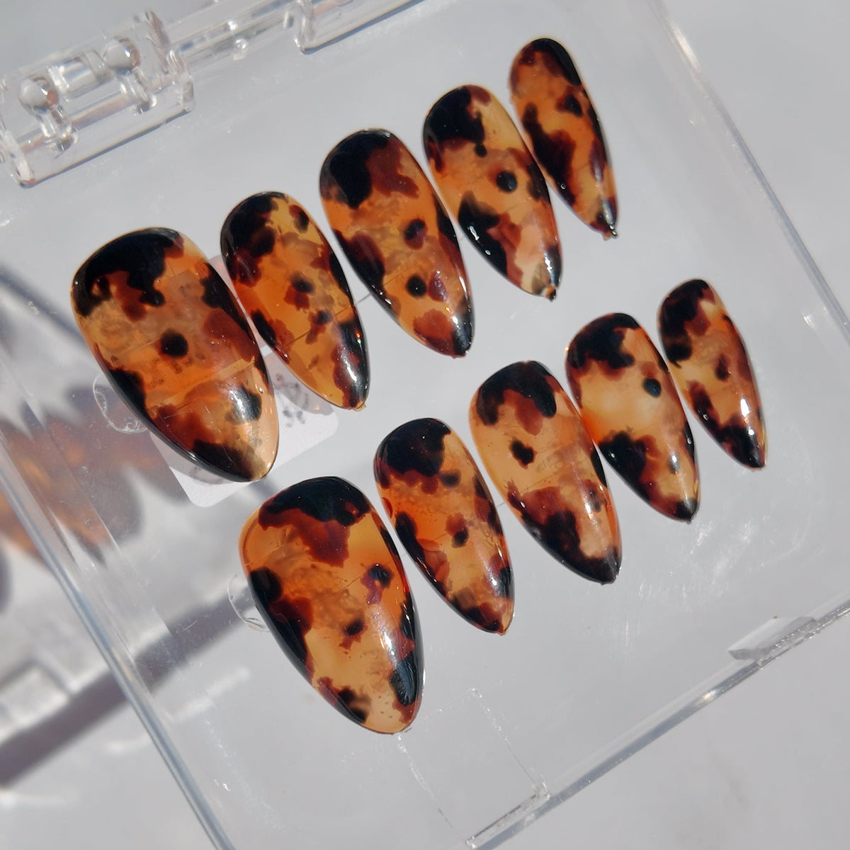 Almond-shaped press-on nails with a glossy tortoiseshell pattern in orange, brown, and black, displayed in a clear plastic case on a light gray surface.