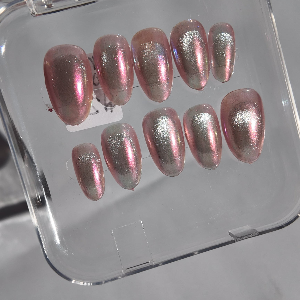 A set of metallic pink press-on nails with a pearlescent shimmer, displayed inside a clear plastic case on a light gray surface.