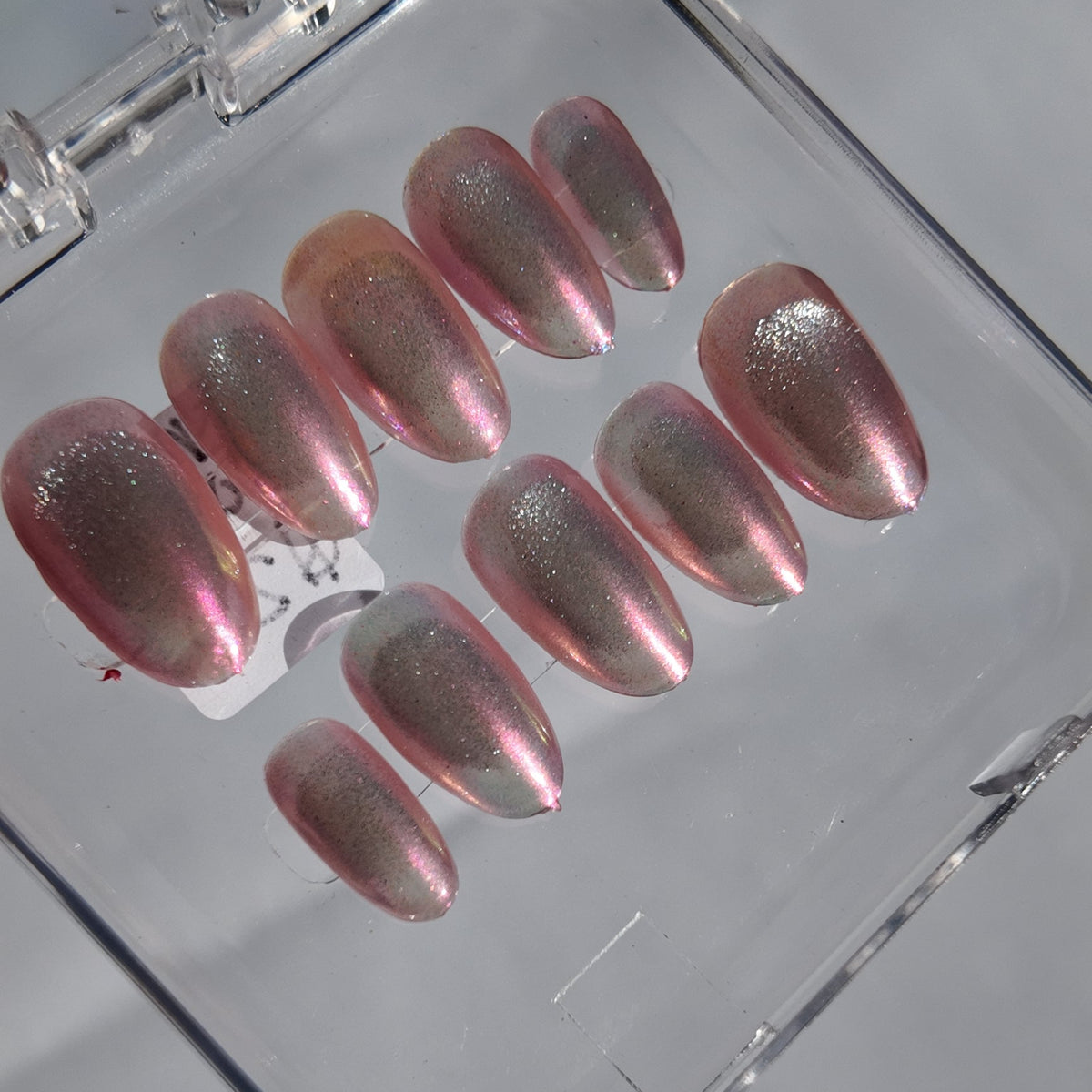 A set of metallic pink press-on nails with a shimmering silver magnetic finish, displayed inside a clear plastic case on a light gray surface.