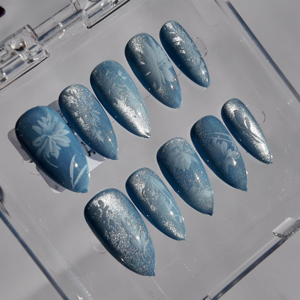 A set of ten press-on nails in a clear plastic case, featuring a light blue base with white glittery patterns and floral designs.