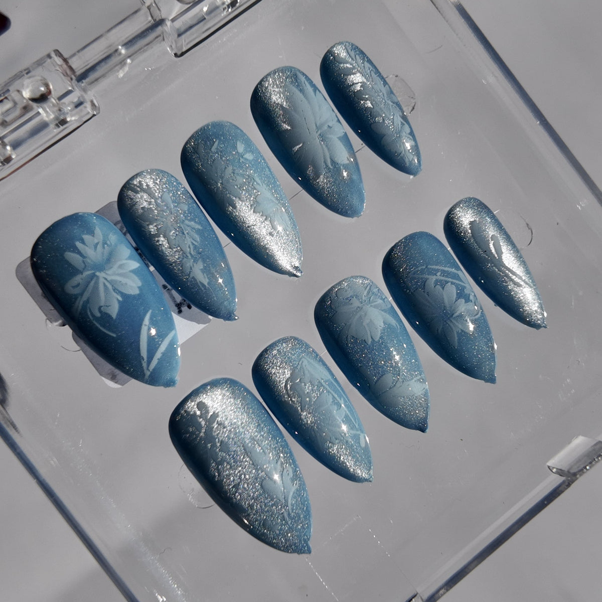 A set of ten press-on nails in a clear plastic case, featuring a light blue base with white glittery patterns and floral designs.