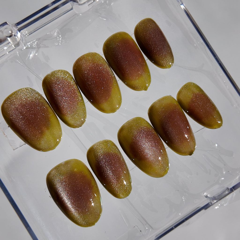 A set of ten false press-on nails with a glossy bronze and olive-green gradient finish, displayed inside a clear plastic storage case on a light gray background.