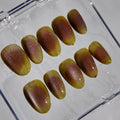 A set of ten false press-on nails with a glossy bronze and olive-green gradient finish, displayed inside a clear plastic storage case on a light gray background.
