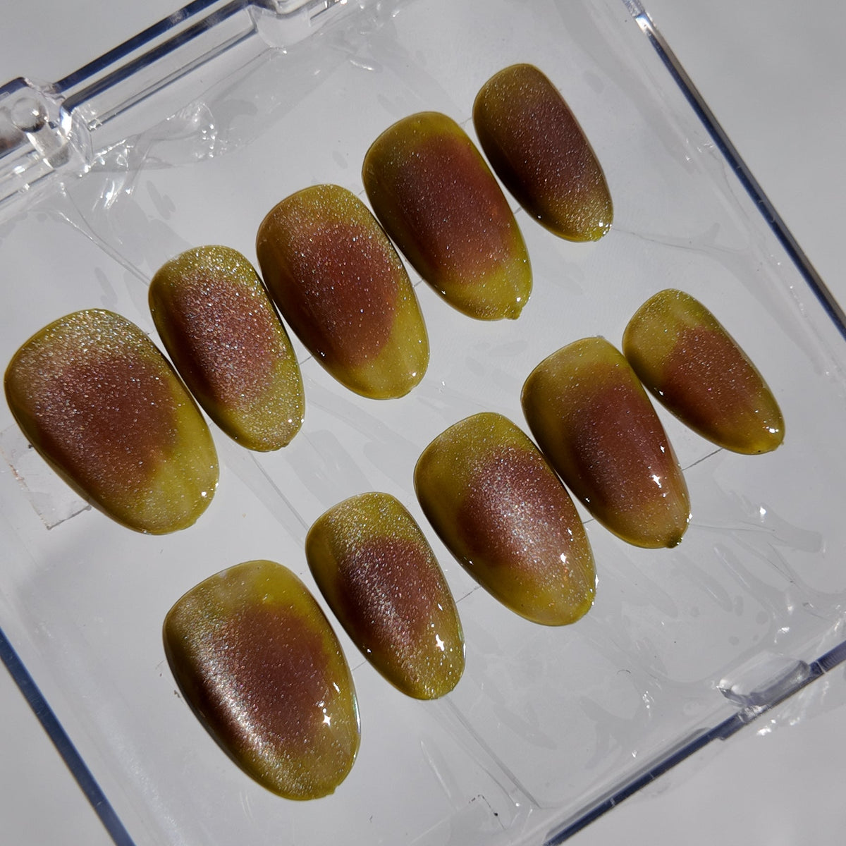A set of ten false press-on nails with a glossy bronze and olive-green gradient finish, displayed inside a clear plastic storage case on a light gray background.