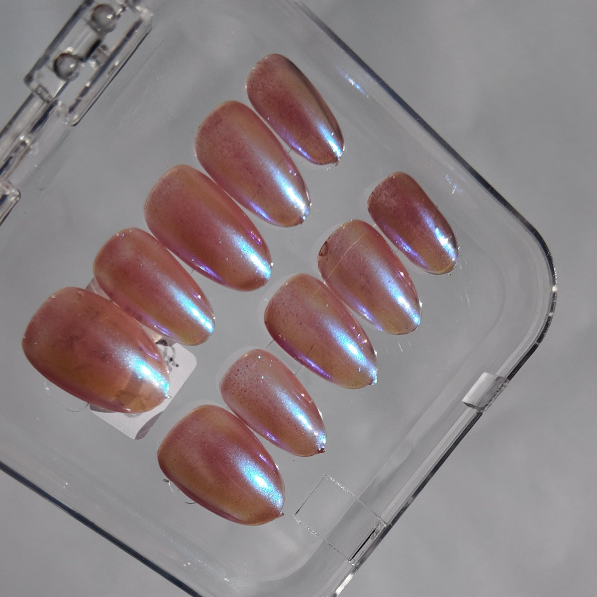 Ten press-on nails with a pearlescent iridescent finish, displayed inside a clear plastic case on a light gray surface.