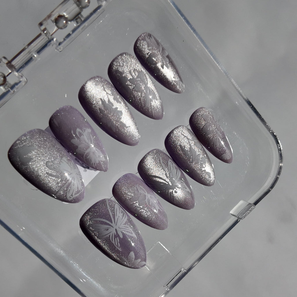 A set of ten press-on nails with a metallic silver glitter finish and white floral patterns, displayed inside a clear plastic case on a light gray background.