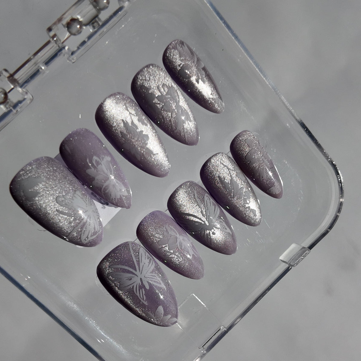 A set of ten press-on nails with a metallic silver glitter finish and white floral patterns, displayed inside a clear plastic case on a light gray background.