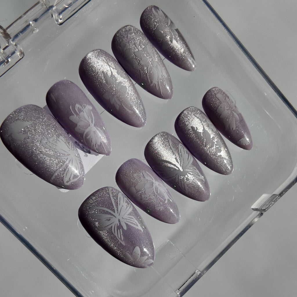 A set of artificial press-on nails in a clear plastic case, featuring a lavender-gray glitter base with white floral patterns.