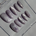 A set of artificial press-on nails in a clear plastic case, featuring a lavender-gray glitter base with white floral patterns.